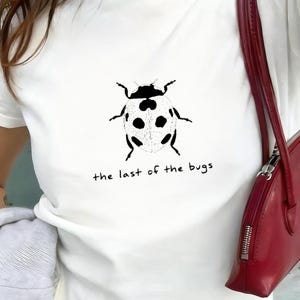 May include: White t-shirt with a black and white ladybug design and the words "the last of the bugs". The top is short-sleeved. A red handbag is visible. The t-shirt is made of a soft material.