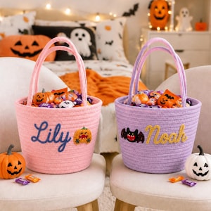 Puede incluir: Dos cestas redondas de cuerda con asas, una rosa y otra morada, llenas de dulces de Halloween. La cesta rosa tiene el nombre Lily en letra azul y una calabaza. La cesta morada tiene el nombre Noah en letra amarilla y un murciélago.