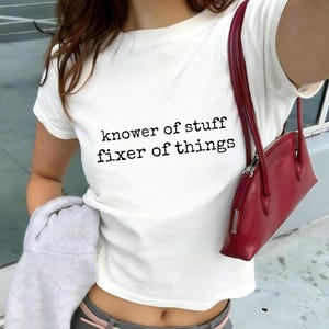 May include: White crew neck t-shirt with the text "knower of stuff fixer of things" in black. A red handbag with a silver zipper is hanging from the shoulder. A gray sweatshirt is held in the arm.