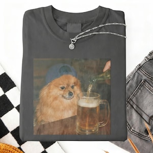 Puede incluir: Camiseta gris oscuro con un meme de un perro Pomerania con una gorra azul, mirando un vaso de cerveza que se está llenando. También es visible un collar plateado con un pequeño colgante. La camiseta está doblada, con un patrón de cuadros blancos y negros.