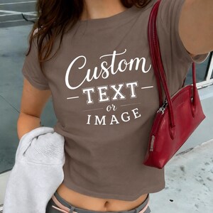 May include: A brown short-sleeved t-shirt featuring white printed text that reads Custom TEXT or IMAGE. The shirt has a crew neckline and is paired with a red shoulder bag and a light gray garment held at the side.