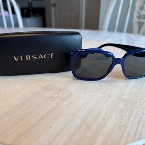 Op de afbeelding: Marineblauwe zonnebril met donkere glazen en een bijpassende etui. De etui heeft de merknaam "VERSACE" in goudkleur. De zonnebril ligt op een lichtgekleurd houten oppervlak.