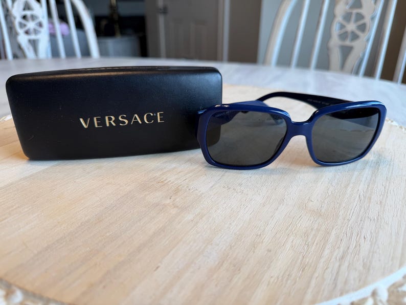 Op de afbeelding: Marineblauwe zonnebril met donkere glazen en een bijpassende etui. De etui heeft de merknaam "VERSACE" in goudkleur. De zonnebril ligt op een lichtgekleurd houten oppervlak.