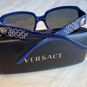 Op de afbeelding: Donkerblauwe zonnebril met een vierkant montuur en grijze glazen. De pootjes hebben een wit geometrisch patroon en een zilveren Medusa-hoofd embleem. De zonnebril ligt op een zwarte etui met de merknaam "VERSACE" in gouden letters.