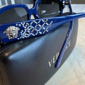Op de afbeelding: Blauwe zonnebril met een kristallen patroon en een zilveren Medusa-hoofd embleem. De zonnebril ligt op een zwarte etui met de merknaam in gouden letters.