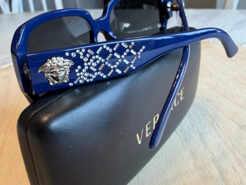 Op de afbeelding: Blauwe zonnebril met een kristallen patroon en een zilveren Medusa-hoofd embleem. De zonnebril ligt op een zwarte etui met de merknaam in gouden letters.