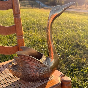 May include: A polished brass swan figurine featuring detailed feather textures on the wings and a long, slender neck. The swan is designed with an open, hollow center, creating a functional vessel shape with a smooth, metallic finish.