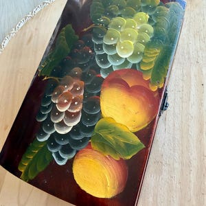 May include: A rectangular, wooden box with a hinged lid. The lid is painted with a still life of grapes and peaches in shades of green, yellow, and red against a dark brown background. The box has a black metal clasp.