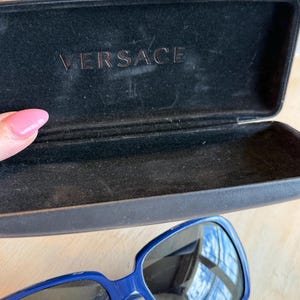 Op de afbeelding: Blauwe zonnebril met een donkere etui. De zonnebril heeft een vierkant montuur en donkere glazen. De etui is zwart en heeft de merknaam "VERSACE" in reli&euml;f aan de binnenkant.