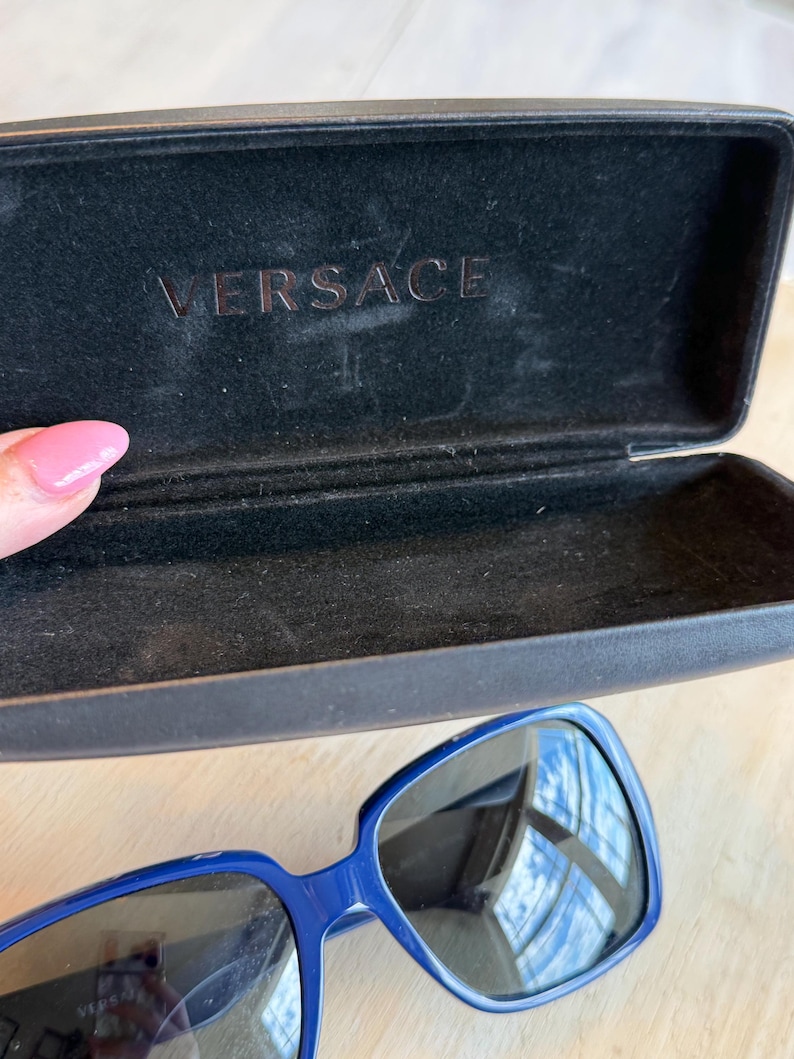 Op de afbeelding: Blauwe zonnebril met een donkere etui. De zonnebril heeft een vierkant montuur en donkere glazen. De etui is zwart en heeft de merknaam "VERSACE" in reli&euml;f aan de binnenkant.