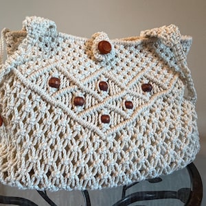 May include: Cream-colored macrame handbag with a geometric design and wooden bead accents. The bag features a woven pattern, a button closure, and a shoulder strap. The bag is a bohemian style.