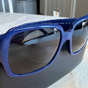 Op de afbeelding: Donkerblauwe Versace zonnebril met donkere glazen. De zonnebril ligt op een zwarte etui. Het montuur is effen, glanzend donkerblauw. De merknaam is zichtbaar op de linker poot.