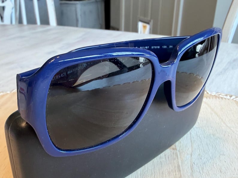 Op de afbeelding: Donkerblauwe Versace zonnebril met donkere glazen. De zonnebril ligt op een zwarte etui. Het montuur is effen, glanzend donkerblauw. De merknaam is zichtbaar op de linker poot.