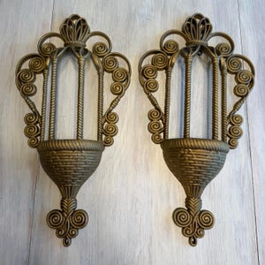 May include: Two ornate, gold-colored wall sconces. Each sconce features a basket-like base, vertical bars, and decorative scrollwork. The sconces are designed to hold small items or candles, adding a touch of vintage elegance to any space.