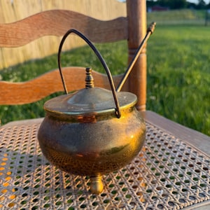 May include: A round brass pot with a lid, featuring a thin metal swing handle and three small feet. The surface of the brass has a warm, aged patina with visible variations in color and texture, and the lid is topped with a small finial.
