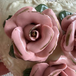 May include: Close-up of a decorative ceramic piece featuring pink roses with gold accents and green leaves. The roses are three-dimensional and set against a cream-colored background with embossed floral patterns. The piece has a glossy finish.