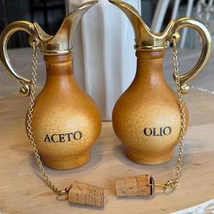 May include: Two ceramic oil and vinegar cruets with gold-colored handles and spouts. The cruets are tan with the words "ACETO" and "OLIO" printed on them. Cork stoppers are attached by a gold chain.