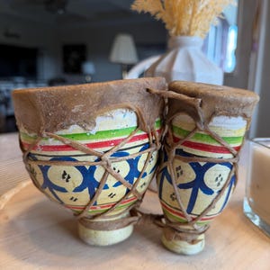 May include: Two small, hand-painted drums with brown leather drumheads. The drums are decorated with blue, red, green, and yellow patterns. The drums are tied together with a natural-colored rope.