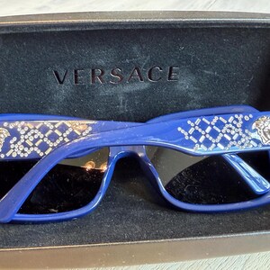 Op de afbeelding: Blauwe Versace zonnebril met een rechthoekig montuur en zwarte glazen. De veren zijn versierd met een zilverkleurig geometrisch patroon en het Versace Medusa-hoofdlogo. De zonnebril zit in een zwart etui met het Versace-logo.