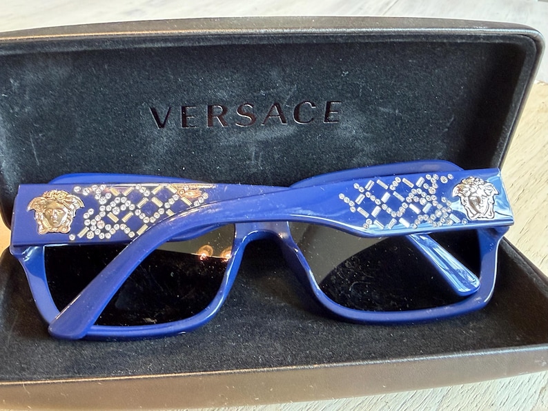 Op de afbeelding: Blauwe Versace zonnebril met een rechthoekig montuur en zwarte glazen. De veren zijn versierd met een zilverkleurig geometrisch patroon en het Versace Medusa-hoofdlogo. De zonnebril zit in een zwart etui met het Versace-logo.