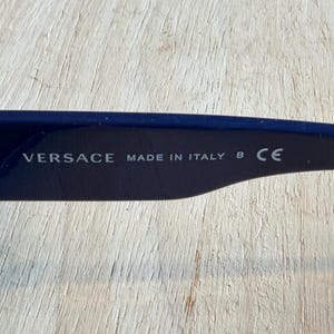 Op de afbeelding: Close-up van de poot van een donkerblauwe zonnebril. De tekst "VERSACE MADE IN ITALY B CE" is in het wit op de binnenkant van de poot gedrukt. De achtergrond is een lichte houtnerf.