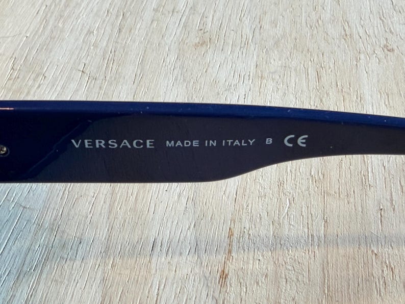 Op de afbeelding: Close-up van de poot van een donkerblauwe zonnebril. De tekst "VERSACE MADE IN ITALY B CE" is in het wit op de binnenkant van de poot gedrukt. De achtergrond is een lichte houtnerf.