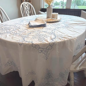 May include: An oval white tablecloth with a blue floral pattern covers a wooden table. A stack of blue napkins, a wooden tray with a candle and vase with dried flowers are on the table. White chairs surround the table.