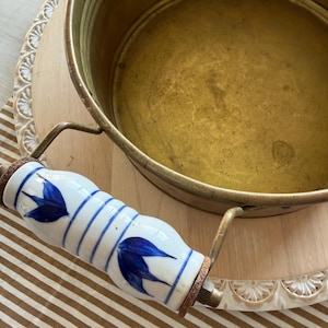 May include: A vintage brass cooking pot with a white handle decorated with blue leaf designs. The pot has a round shape and is sitting on a wooden surface with a striped brown and white fabric.