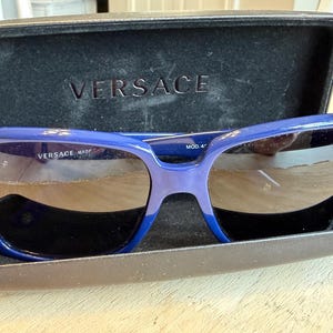 Op de afbeelding: Blauwe Versace zonnebril met donkere glazen in een open zwarte etui. De merknaam is zichtbaar op de etui en de zonnebril. De zonnebril heeft een dik, rechthoekig montuur met een verloop van blauwtinten.