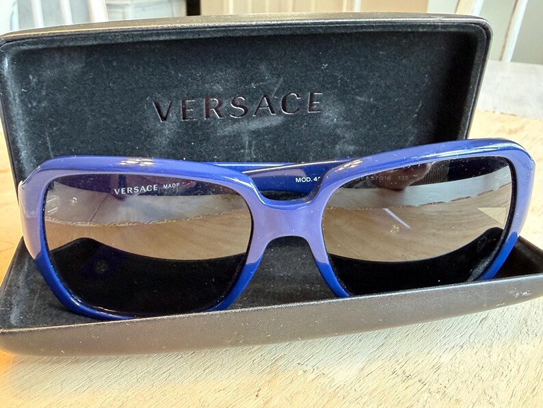 Op de afbeelding: Blauwe Versace zonnebril met donkere glazen in een open zwarte etui. De merknaam is zichtbaar op de etui en de zonnebril. De zonnebril heeft een dik, rechthoekig montuur met een verloop van blauwtinten.