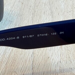 Op de afbeelding: Close-up van de binnenarm van een marineblauwe zonnebril. De tekst "MOD. 4204-B 911/87 57▢16 135 3N" is in het wit op de zwarte arm gedrukt. De zonnebril ligt op een licht houten oppervlak.