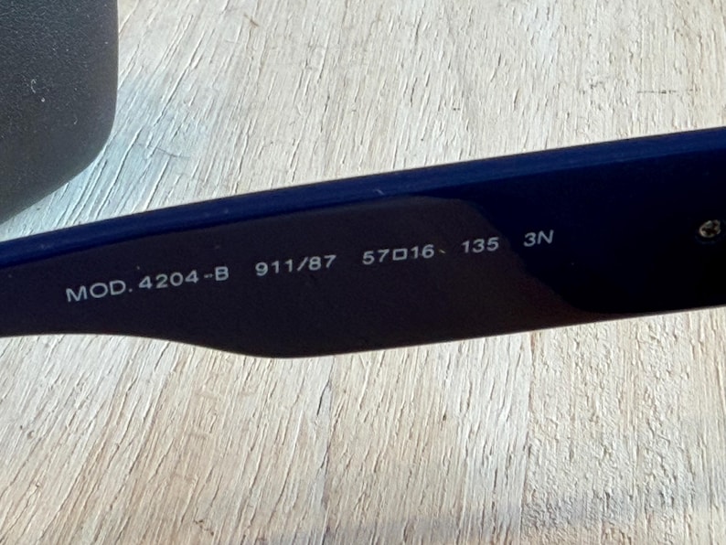 Op de afbeelding: Close-up van de binnenarm van een marineblauwe zonnebril. De tekst "MOD. 4204-B 911/87 57▢16 135 3N" is in het wit op de zwarte arm gedrukt. De zonnebril ligt op een licht houten oppervlak.