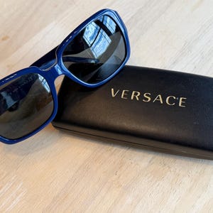 Op de afbeelding: Blauwe zonnebril met donkere glazen op een zwarte etui. De zonnebril heeft een rechthoekig montuur. Op de etui staat in gouden letters "VERSACE". De zonnebril en etui liggen op een licht houten oppervlak.