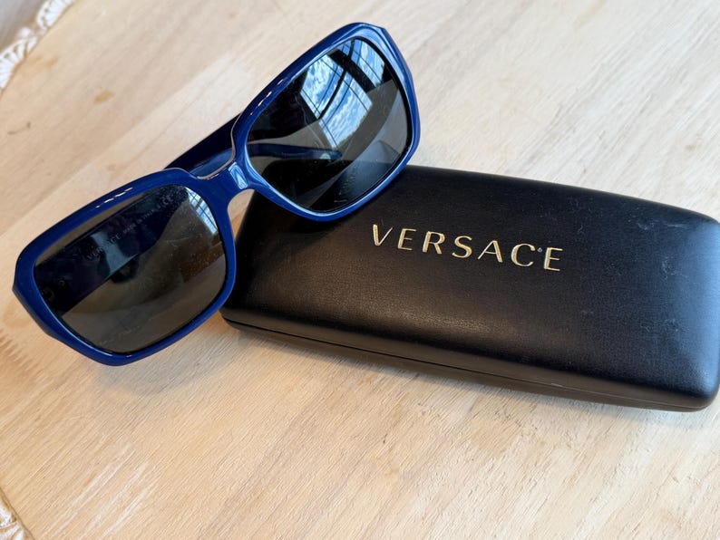 Op de afbeelding: Blauwe zonnebril met donkere glazen op een zwarte etui. De zonnebril heeft een rechthoekig montuur. Op de etui staat in gouden letters "VERSACE". De zonnebril en etui liggen op een licht houten oppervlak.