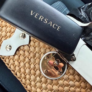 Op de afbeelding: Een zwarte Versace zonnebril etui met gouden letters ligt op een geweven strooien handtas. De handtas heeft witte lederen details en een goudkleurige sluiting met het Michael Kors logo.