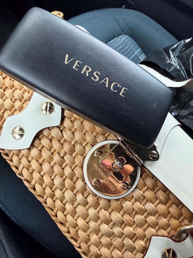 Op de afbeelding: Een zwarte Versace zonnebril etui met gouden letters ligt op een geweven strooien handtas. De handtas heeft witte lederen details en een goudkleurige sluiting met het Michael Kors logo.