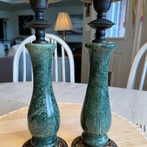 May include: Two antique candle holders. The bases are ornate, with a dark bronze finish. The stems are a green marble, and the tops are a bronze color. The candle holders are on a light-colored wooden table.