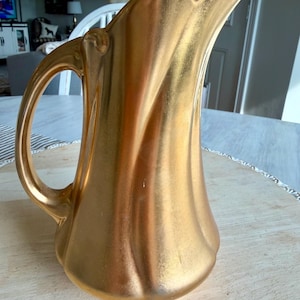 May include: A gold-colored pitcher with a curved spout and a rounded body. The pitcher has a handle on the side and is made of a shiny, metallic material. The pitcher is sitting on a light-colored surface.