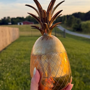 May include: A metallic gold-toned pineapple-shaped container with a textured diamond-patterned body and a pointed leafy top. The object features a reflective surface that catches the light, showcasing a distinct segmented design and a removable lid.