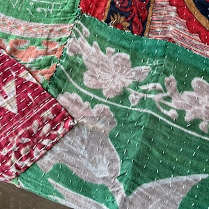 May include: Close-up of a patchwork textile with various patterns and colors. The textile features sections of red, green, and patterned fabrics stitched together with white thread. Floral and geometric designs are visible.