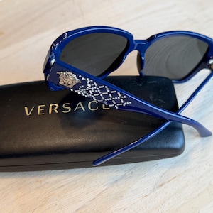 Op de afbeelding: Blauwe zonnebril met donkere glazen rust op een zwarte etui met het gouden woord "VERSACE". De zonnebril heeft een decoratief patroon met zilverkleurige accenten op de veren. Het montuur is glanzend, diepblauw.