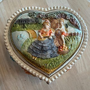 May include: Heart-shaped decorative box with a raised relief scene. The box features a woman playing a guitar, a cherub, and a basket of fruit. The box is cream-colored with a beaded border and a detailed landscape.