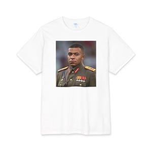 Pode incluir: Uma t-shirt branca de manga curta com uma impressão gráfica retangular centrada de uma pessoa com cabelo curto e escuro, vestindo um uniforme militar castanho com dragonas douradas, abas vermelhas e várias medalhas prateadas no peito.