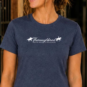 May include: A heathered navy blue short-sleeve t-shirt featuring a white graphic print on the chest. The design includes silhouettes of horses and riders with the text Thoroughbred FROM THE TRACK TO THE CENTERLINE in a script and sans-serif font.