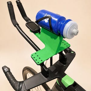 Könnte beinhalten: Ein Fahrrad-Cockpit mit schwarzen Aero-Lenkern und schwarzem Lenkerband, einer hellgrünen, mittig angebrachten Halterung, einer blauen Trinkflasche in einem schwarzen Flaschenhalter sowie einem kleinen, schwarzen digitalen Fahrradcomputer.