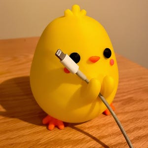 May include: A yellow chick-shaped cable organizer with black eyes, orange beak, and pink cheeks. The chick holds a white and gray charging cable. The organizer is on a wooden surface.