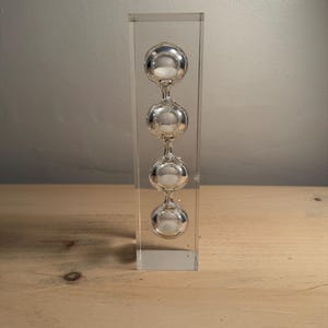 May include: A clear rectangular acrylic block containing a vertical series of four interconnected glass spheres. Each sphere features a small white circular element at its centre, creating a stacked, linear arrangement within the transparent casing.