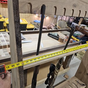 K&ouml;nnte beinhalten: Ein h&ouml;lzernes Gewehrregal mit nat&uuml;rlicher Kante, etwa 127 cm breit. Das Regal hat geformte Schlitze zur Aufnahme von Schusswaffen und zeigt zwei Gewehre. Ein Ma&szlig;band wird an das Regal gehalten.