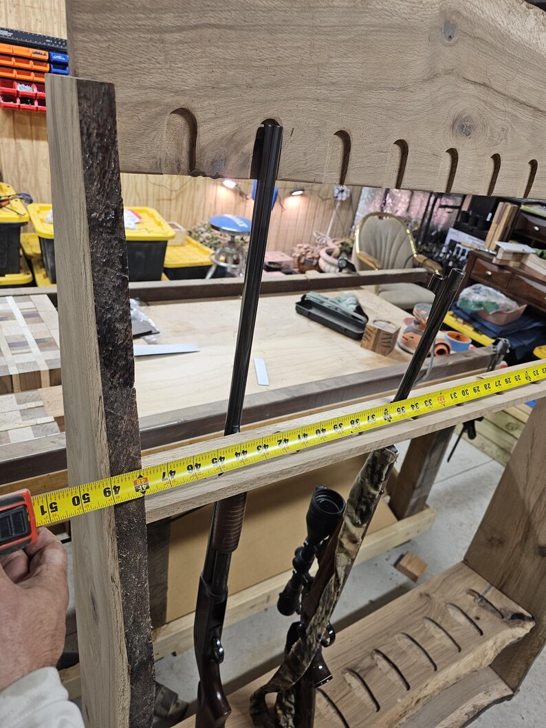 K&ouml;nnte beinhalten: Ein h&ouml;lzernes Gewehrregal mit nat&uuml;rlicher Kante, etwa 127 cm breit. Das Regal hat geformte Schlitze zur Aufnahme von Schusswaffen und zeigt zwei Gewehre. Ein Ma&szlig;band wird an das Regal gehalten.