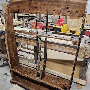 K&ouml;nnte beinhalten: Ein rustikaler Gewehrst&auml;nder aus Holz mit nat&uuml;rlicher Kante. Der St&auml;nder, mit dunkelbraunem Finish, hat Schlitze f&uuml;r mehrere Schusswaffen, wobei zwei Gewehre ausgestellt sind. Das Design hebt die nat&uuml;rliche Maserung und Kante des Holzes hervor.